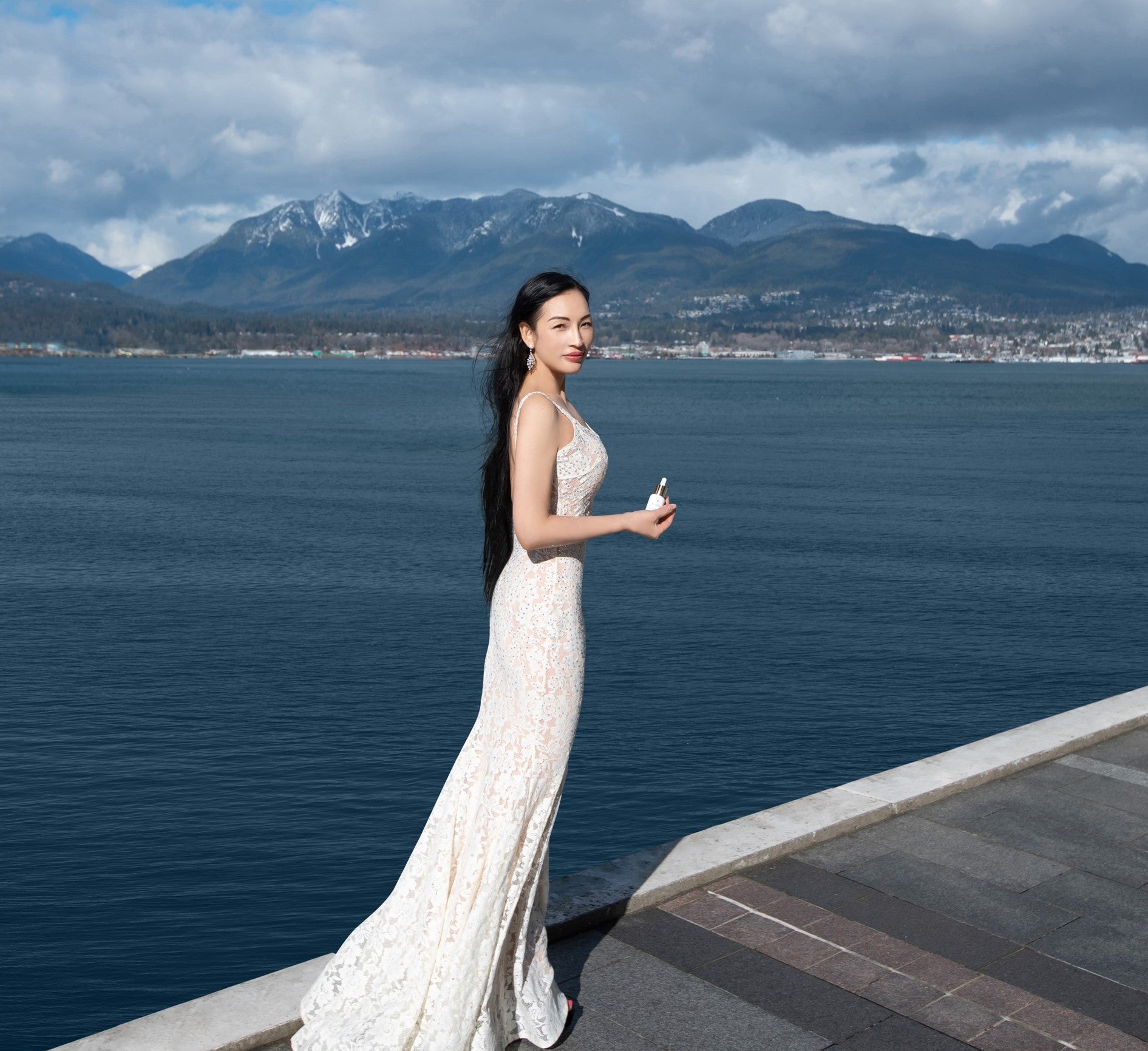 Woman in a white dress standing by a waterfront with mountains in the background